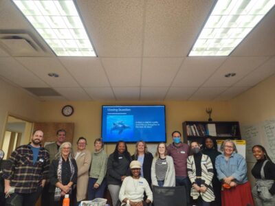 A group of 11 people who participated in the Disability Rights North Carolina Structured Negotiation training, with Lainey Feingold and Barry Taylor who conducted the training. They are standing in front of a slide with an image of dolphins