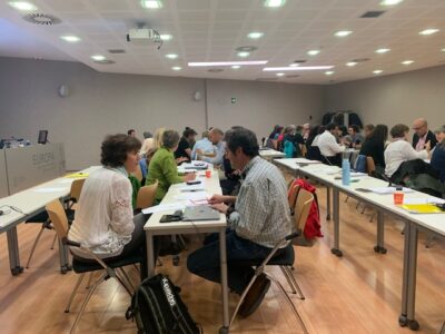 Around 25 people gathered around two long tabled working together at a Structured Negotiation workshop in Spain