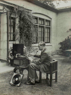 Black and white photo of a pipe-smoking man in a suit sitting in a garden while putting a new record on a phonograph player.
