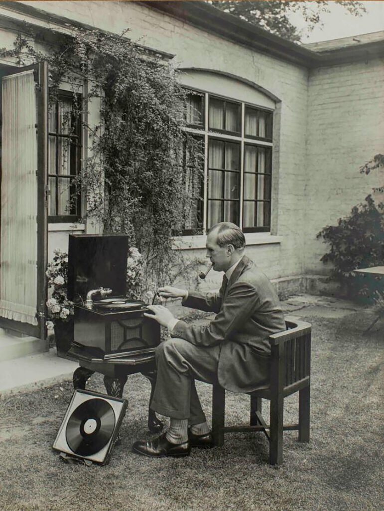 Black and white photo of a pipe-smoking man in a suit sitting in a garden while putting a new record on a phonograph player.