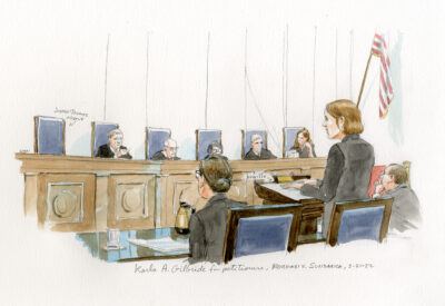 A courtroom sketch of Karla Gilbride, a white woman, standing at the lectern giving an argument to the U.S. Supreme Court justices. The sketch shows her hands on her Braille notes with an arrow indicating “Braille.” The sketch also shows Karla’s co-counsel taking handwritten notes, her opposing counsel listening, and Justice Thomas’s empty chair among those of the other seated justices, indicating that he was not present for the argument. There are American flags in the background.