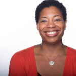 Reginé Gilbert is a Black woman with short curly hair smiling at the camera wearing an orange cardigan and a pendant necklace.