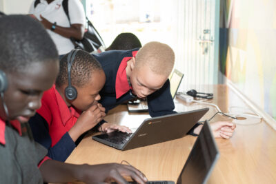 Three visually impaired Kenyan students sit side by side at a desk using laptops in a brightly lit digital lab. Two learners, one of whom has albinism, are closely focused on a shared Chromebook, wearing headphones and engaging in a computer-based activity. Another student is in the foreground, also using a laptop. A teacher stands in the background near the door.