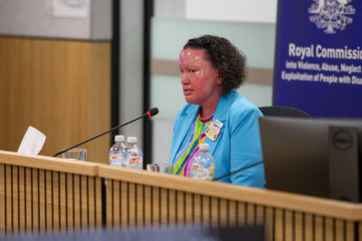 Carly Findlay, a woman with red skin and short dark curly hair, wearing a blue suit and a rainbow t-shirt, sitting at a desk, testifying at the Disability Royal Commission.