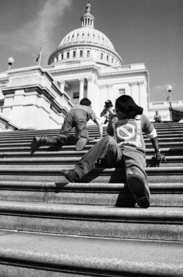 Black and white photo taken from the bottom of a large set of concrete steps. It captures two people with disabilities crawling up the steps of the US Capitol with the Capitol dome towering above them. The man is crawling on his hands and knees. The woman is crawling up backwards, resting on her rear on each step. The woman is Black, the man is white.